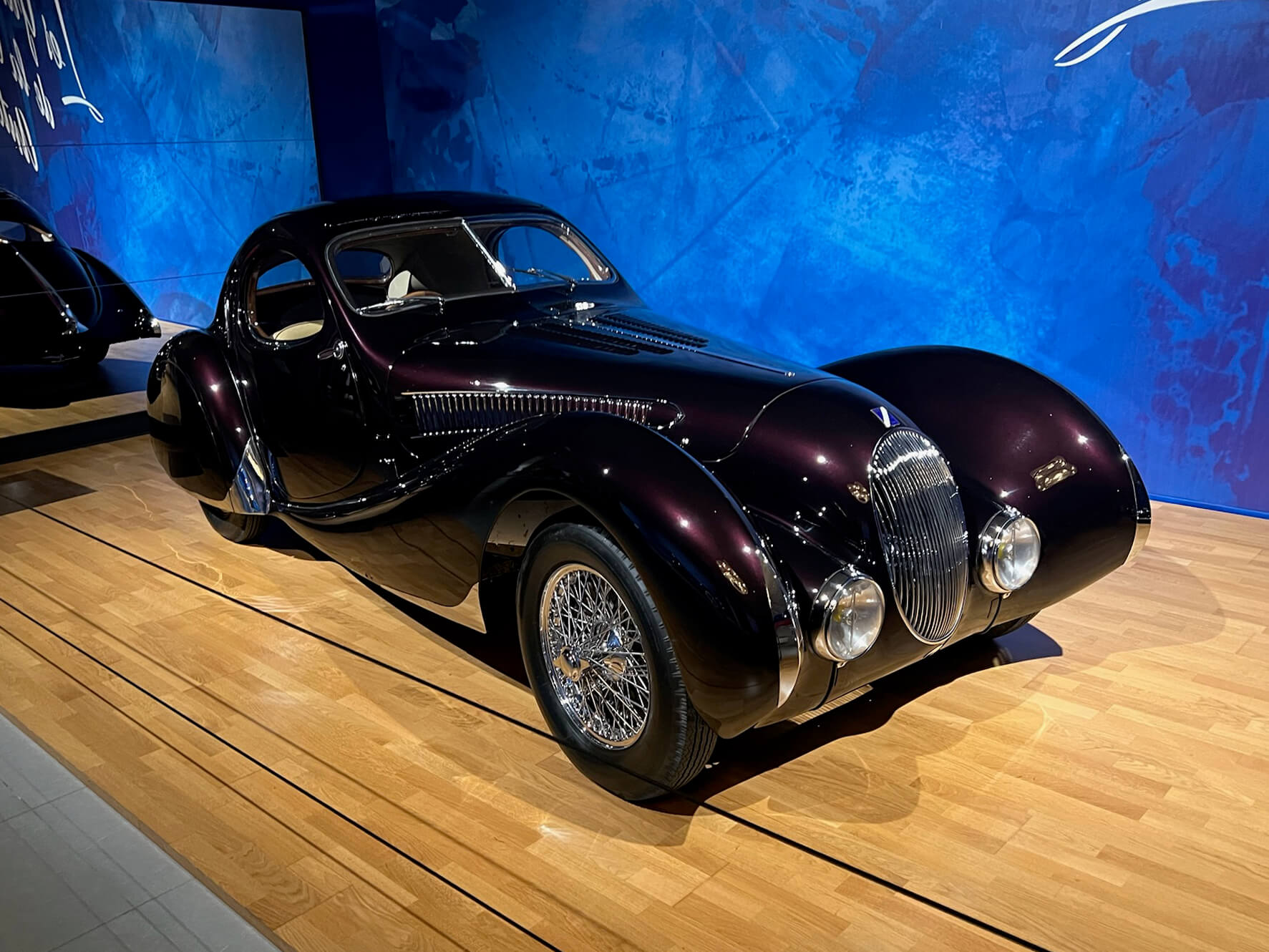 Dark coachbuilt Bugatti Type 57SC Atlantic displayed at the Louwman Museum with a dramatic blue backdrop and polished chrome grille
