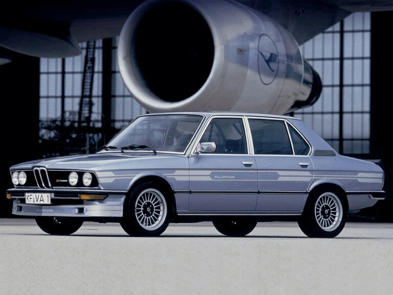 Silver Alpina-modified BMW sedan photographed in profile with an aircraft turbine backdrop, featuring Alpina side graphics and period multi-spoke wheels